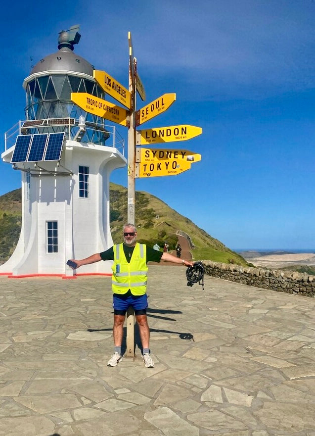 A man wearing a bright yellow safety vest and blue shorts stands in front of a white lighthouse and directional signpost on a stone path under a blue sky.