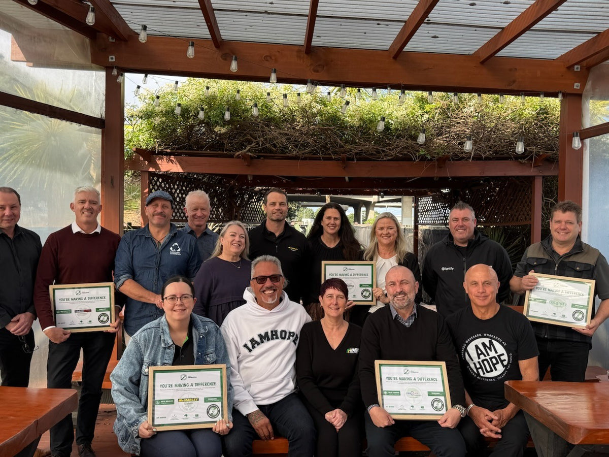 A group of people posing together under a wooden pergola, each holding a framed certificate. The setting appears to be an outdoor covered patio or pavilion with greenery and hanging lights in the background.