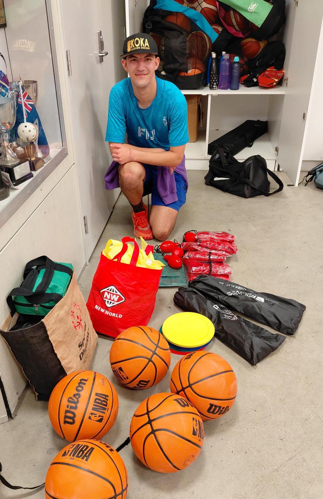A young man in a blue shirt and purple shorts kneels on a concrete floor surrounded by basketballs, bags, and sports gear in a locker room setting.